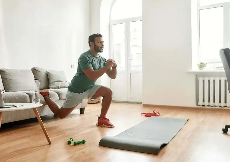 رجل يقوم بتمارين مقاومة في المنزل باستخدام وزن الجسم، يؤدي تمرين القرفصاء البلغاري مع وجود لابتوب بجانبه