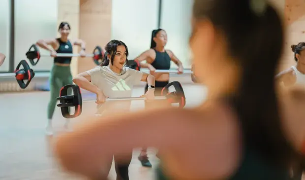 Women participating in a Les Mills BODYPUMP class at GymNation, performing upright row exercises with barbells inside a bright studio